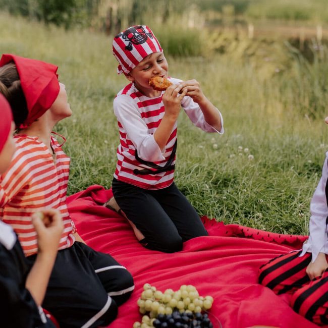 Als Piraten verkleidete Kinder sitzen auf einer Picknickdecke und essen während eines Kindergeburtstags.
