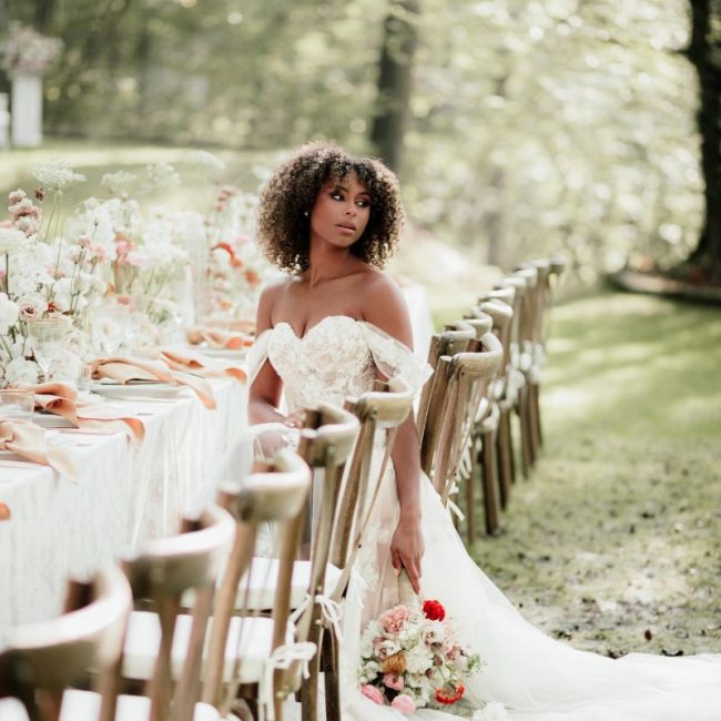 Eine Braut mit einem Strauß in der Hand sitzt an einer Hochzeitstafel.